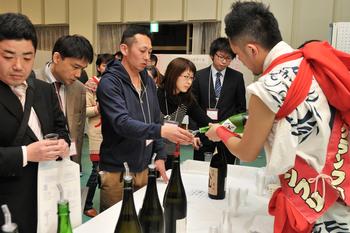 長机の上に銘酒の瓶が並び、法被を着た男性が机の前に並んだ人に酒を提供している写真