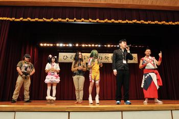 体育館のステージ上でマイクを持っている男性と、コスプレをしている5人の男女の写真