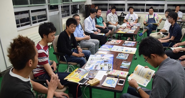 沢山の雑誌やチラシが広げられた机を取り囲んで座る男性たちの写真