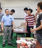 雑誌やチラシが広げられた机の周りに立つ男性たちの写真