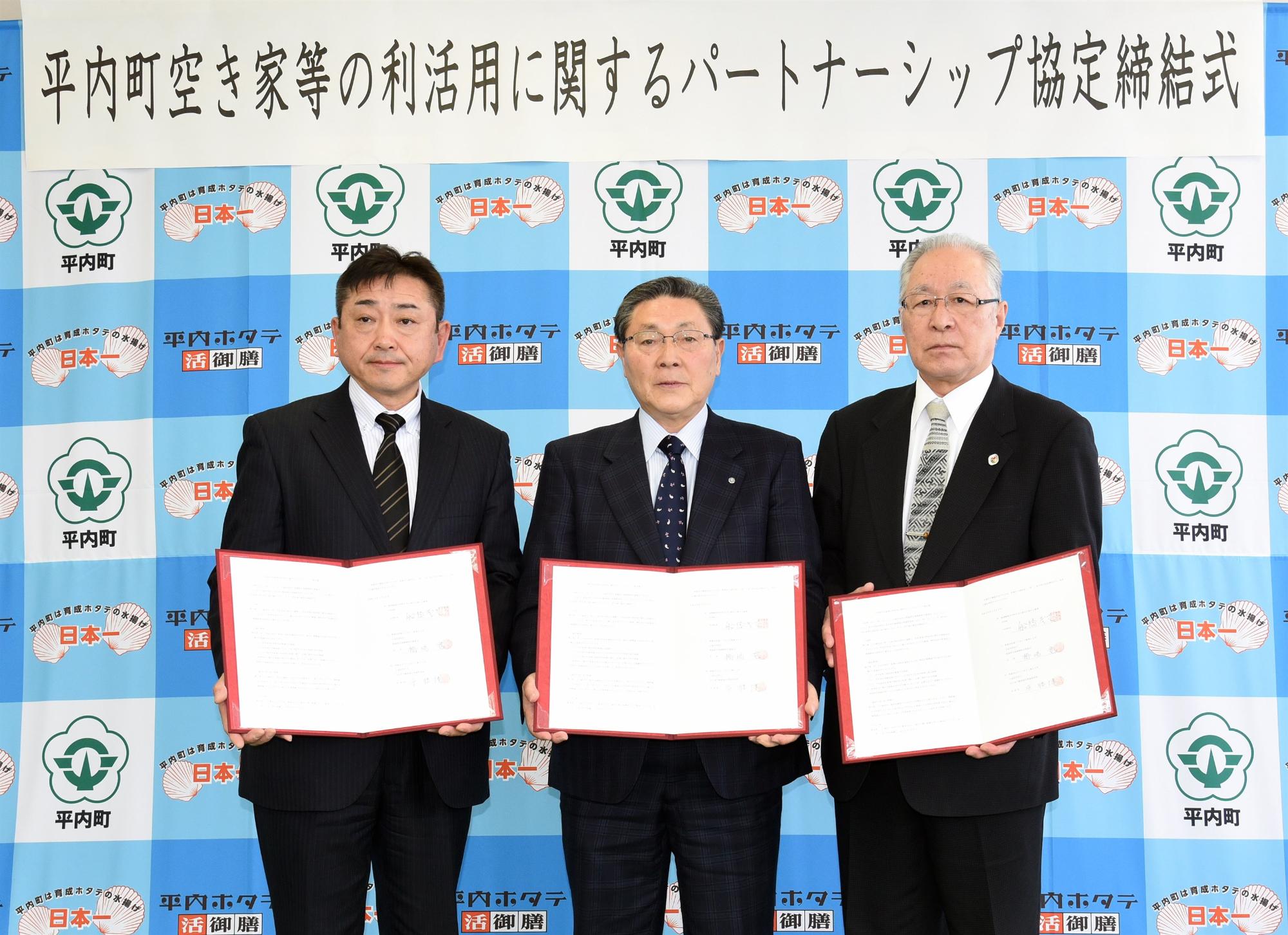 3人のスーツ姿の男性が、インタビューボードの前で証書を手に持って並んでいる写真