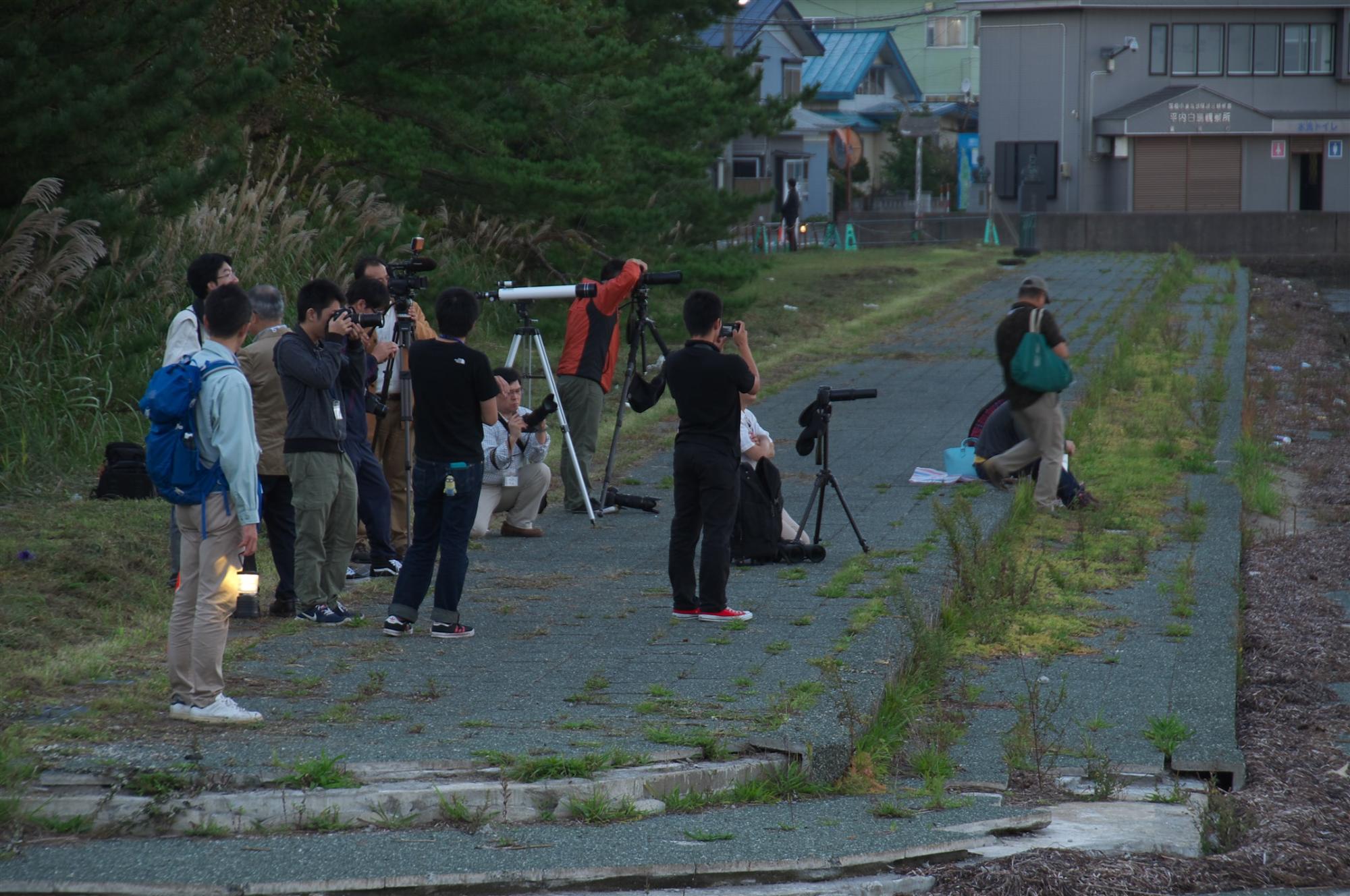 薄暗い時間の道路に望遠鏡を置いて空を眺める人たちの写真