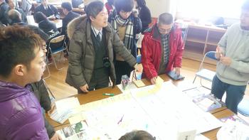 机に広げられた資料を基に話し合いをする男性たちの写真