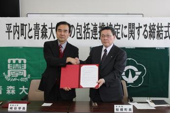 二つの紋章の旗が貼られた壁の前で、スーツ姿の2人の男性が握手しながら証書を見せている写真