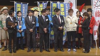 スーツ姿の男性4人と、その他大勢の人がのぼり旗の前に並んでいる写真