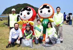 赤の服と緑の服を着たマスコットキャラの周りに6人が囲むように立ったり座ったりしている写真