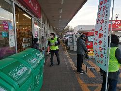 店の入り口前にて男性が女性買い物客に啓発物品を渡している写真
