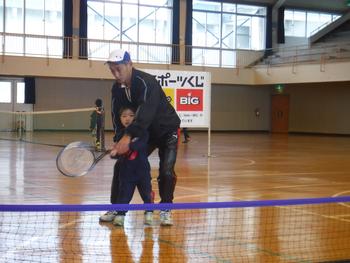 体育館の中で、キャップを被ったジャージの男性が、小さな子どもの後ろに立ってラケットの持ち方を教えている写真