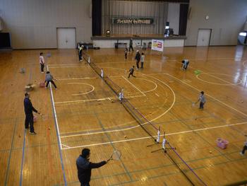 体育館のコート床に貼られたネットを挟み、大人と子どもの対面でテニスをしている姿を上から見た写真