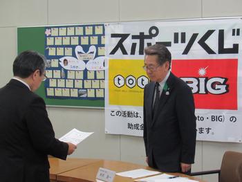 「スポーツくじ」と書かれたパネルの壁を背に、スーツを着たメガネの男性が立ち、目の前の男性から書類の説明を聞いている写真