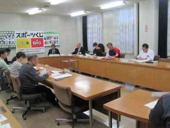 壁に掲示板とスポーツくじのパネルが飾られた会議室に、楕円形に並べられた机でスーツを着た人たちが会議している写真