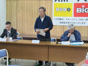 3人の男性が長机の席に並び、中央の人がプリントを持って席を立ち、喋っている写真