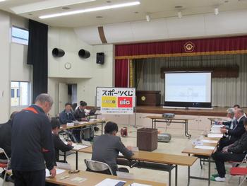 ステージがある会場にスクリーンが下げられ、「スポーツくじ」と書かれたパネルを前に、机を並べて会議している人たちの写真