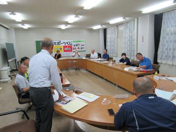 細長い楕円形の会議机がある会議室で、1人の男性が立ちながら会議をしている人たちの写真