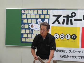 掲示板と布のポスターが掛けられた壁を背に、プリントを持って説明をしている眼鏡の男性の写真