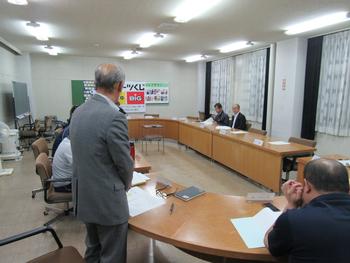 楕円型に配置された机がある会議室で、グレーのスーツの男性が立ち上がって話している会議中の写真