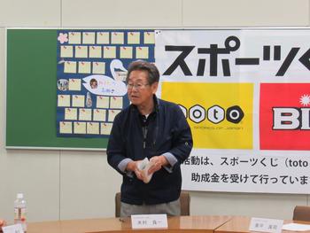 緑色の掲示板と布が飾られた壁の前に立ち、机の前でプリントを持って喋っている眼鏡の男性の写真