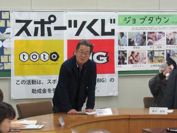 「スポーツくじ」と書かれた布とポスターが飾られた部屋の壁の前に立ち、机に手をついて喋っている眼鏡の男性の写真