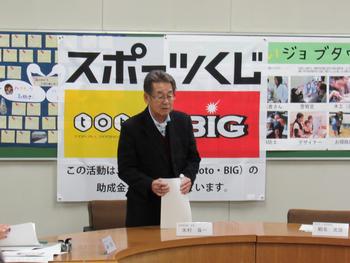 「スポーツくじ」と書かれた布と掲示板がかけられた壁を背に、プリントを手にスピーチしている眼鏡の男性の写真