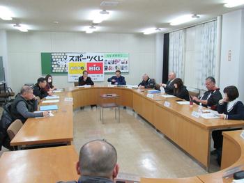 会議室で、楕円状に並べた長机に座った人たちが会議をしている写真
