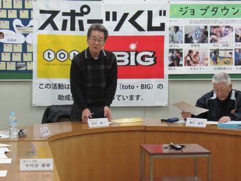 「スポーツくじと」書かれた掲示物の前で話をする、チェックのセーターを着た男性の写真