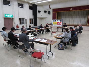 スポーツくじと書かれたホワイトボードの前で四角に机を並べ会議をしている男女12名の写真