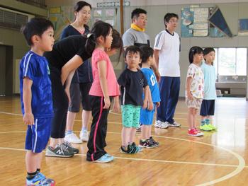 運動靴を履いてTシャツを着た6人の小さな子どもたちと、4人の大人が体育館に立っている写真