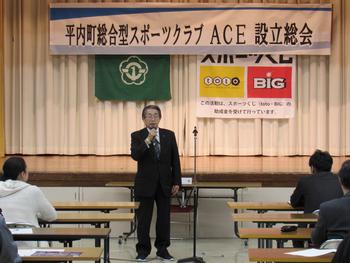 「平内町総合型スポーツクラブ ACE 設立総会」と書かれた垂れ幕の前で話をするスーツの男性の写真