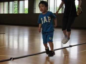 青いTシャツと短パンを着た男の子が、後ろの大人と一緒に体育館のコート上でジャンプしている写真