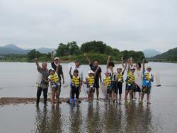 川の中で黄色いライフジャケットを着た子供たちのと私服の大人たちが手を挙げている写真