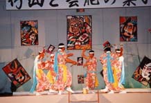 複数のポスターが張られた舞台で、衣装を着た人たちが輪になって踊っている写真