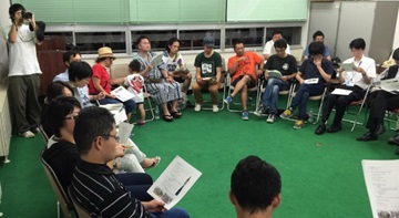緑の床に円形に並べた椅子に資料を見ながら座る人たちの写真