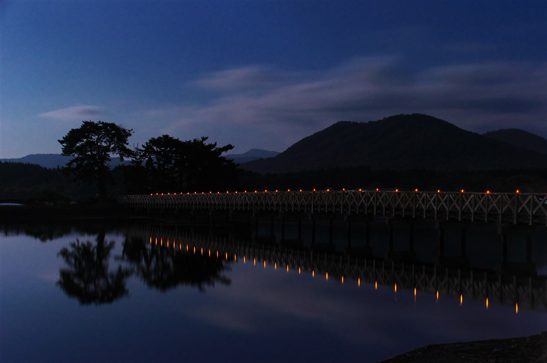 夜の水面に写る木と山と橋の明かりの写真