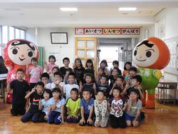 児童たちと赤い服と緑の服を着たマスコットキャラが並んで集合写真を撮っている写真
